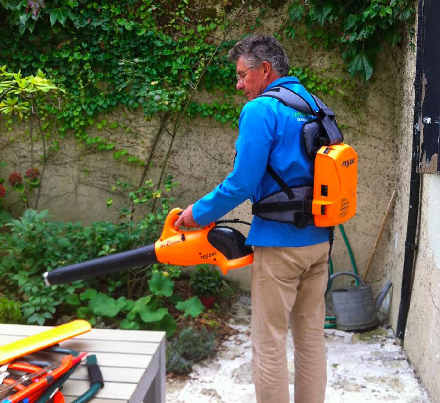 Un homme en veste bleue souffle des feuilles avec un souffleur orange sur batterie porté dans le dos, dans une cour avec un mur couvert de lierre.