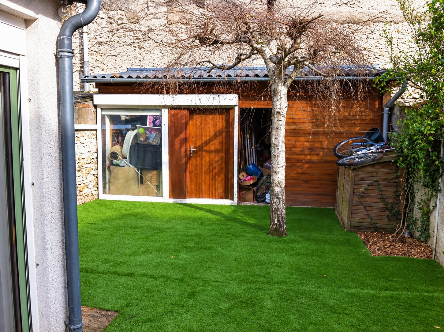 Une petite cour avec une pelouse synthétique d'un vert uniforme, un bouleau et un abri de jardin en bois au fond.