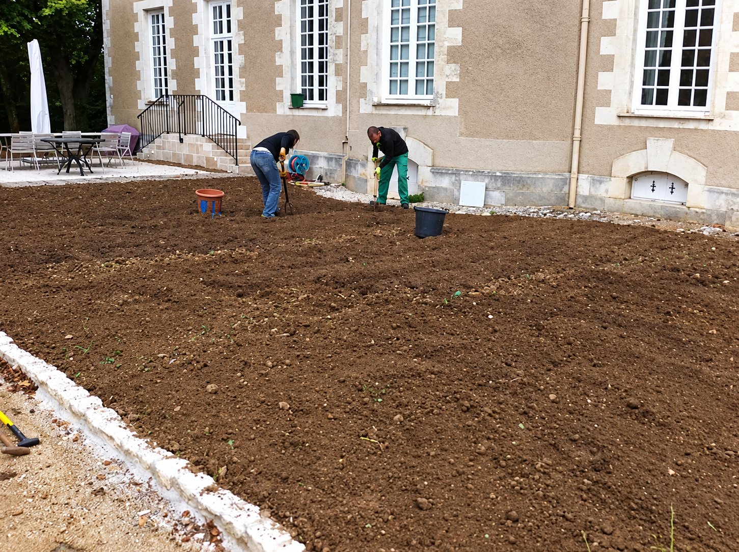 Deux jardiniers travaillent la terre brune d'un futur gazon devant une belle demeure en pierre claire, avec une terrasse et des chaises à gauche.