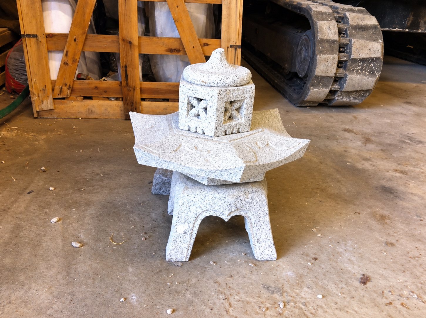 Une petite lanterne japonaise en pierre claire, avec toit recourbé et ouvertures ciselées, posée sur un sol en béton devant une caisse en bois et la chenille d'une machine.