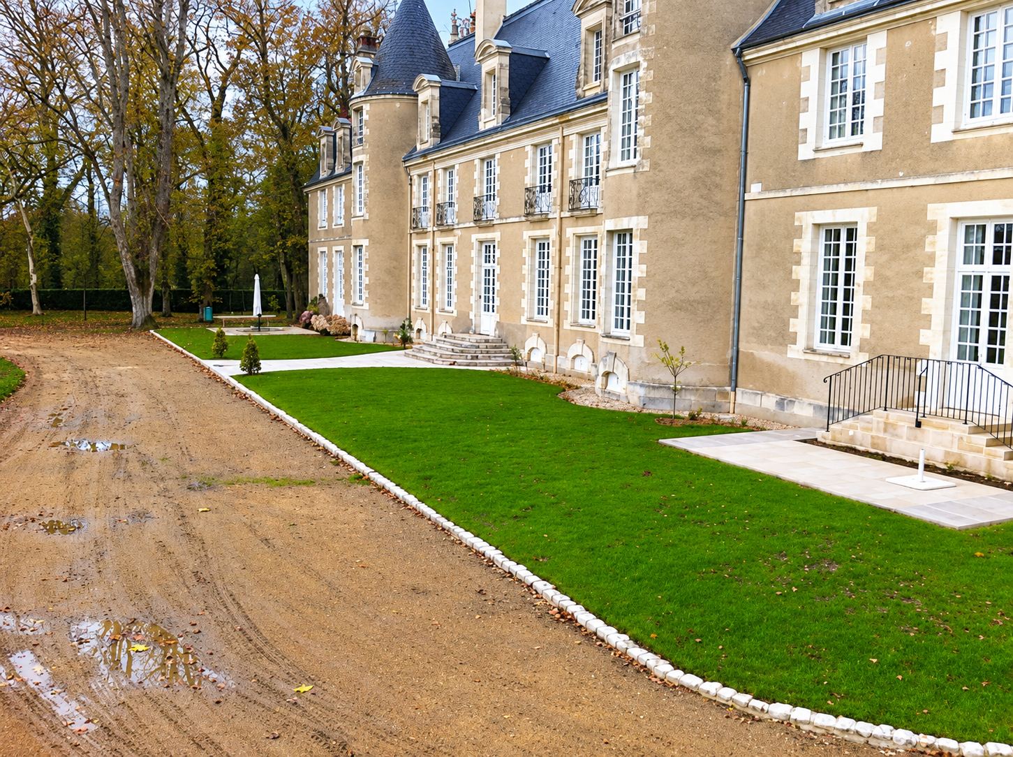 Une pelouse fraîchement posée bordée de pierres blanches longe la façade d'un château en pierre, à côté d'une allée de terre encore en travaux.