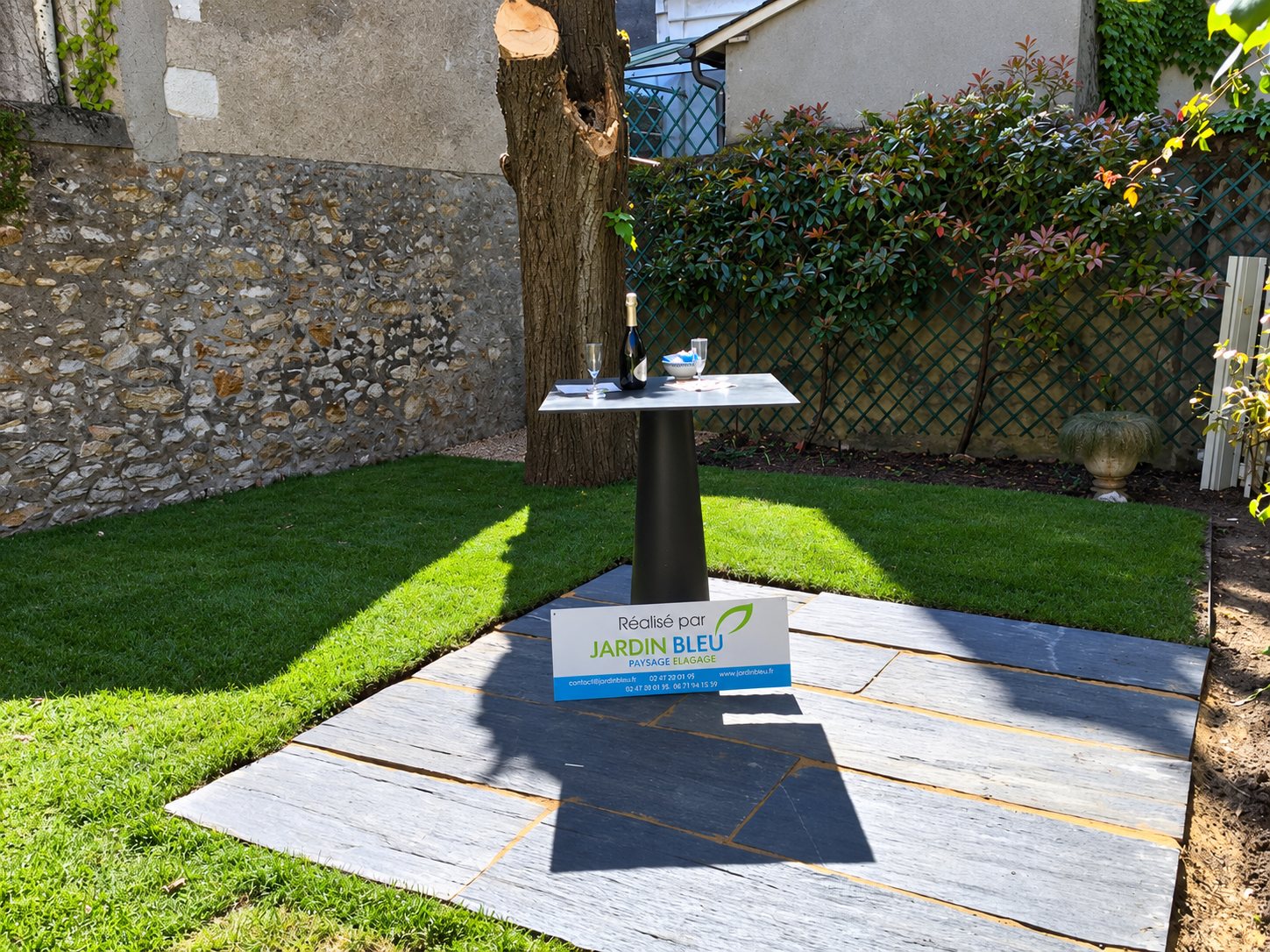 Jardin avec pelouse neuve et terrasse en dalles d'ardoise, table haute et panneau Réalisé par Jardin Bleu devant un mur de pierre.