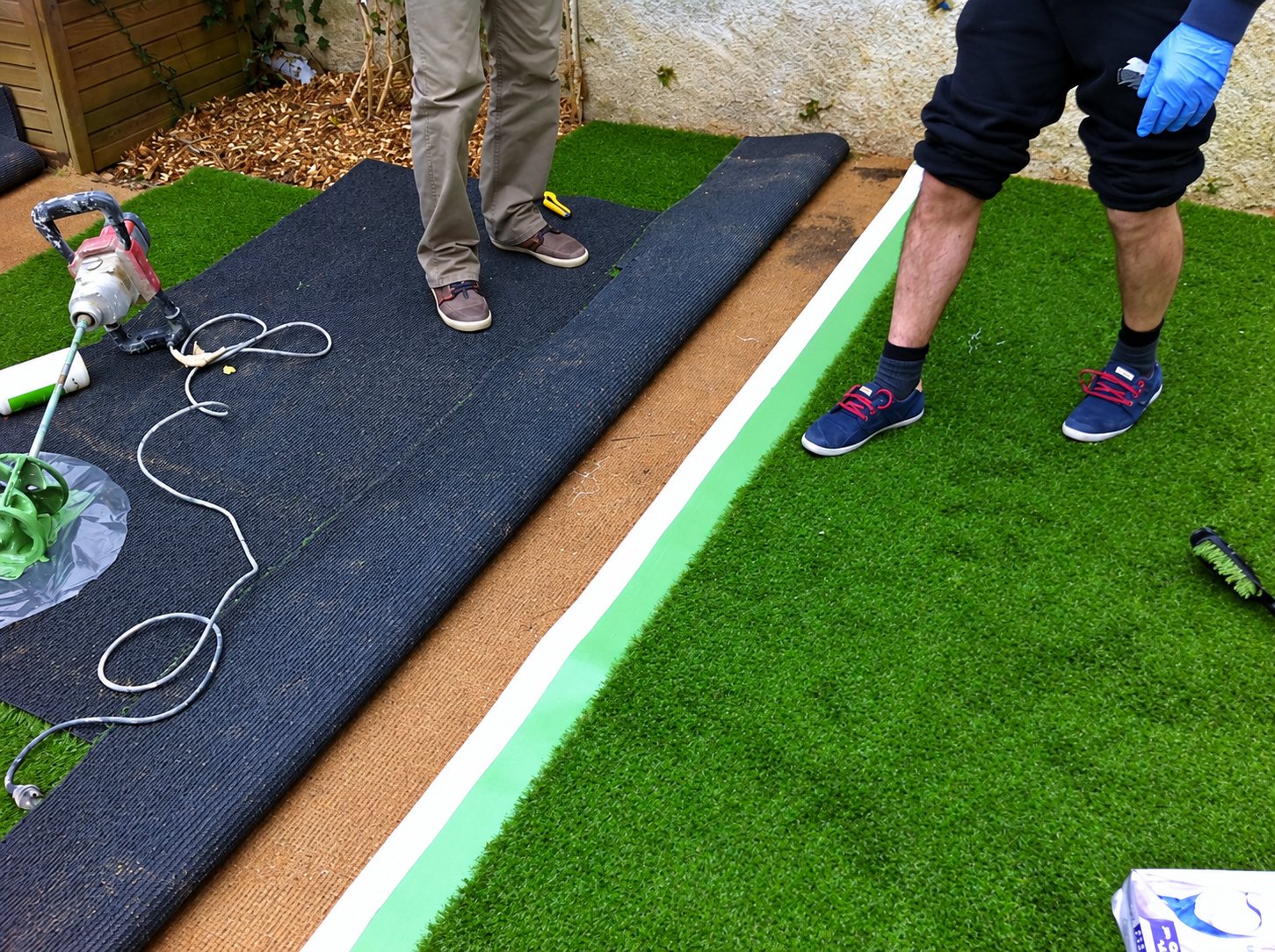 Deux ouvriers posent du gazon synthétique dans un jardin, avec un rouleau de gazon déroulé, un bandeau de jonction vert et un mélangeur de colle au sol.