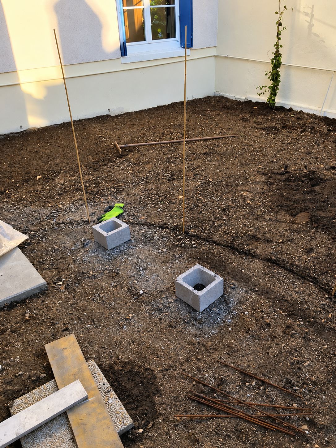Chantier d'aménagement extérieur avec terre nivelée, deux parpaings creux, des piquets en bambou et un râteau devant la façade d'une maison.