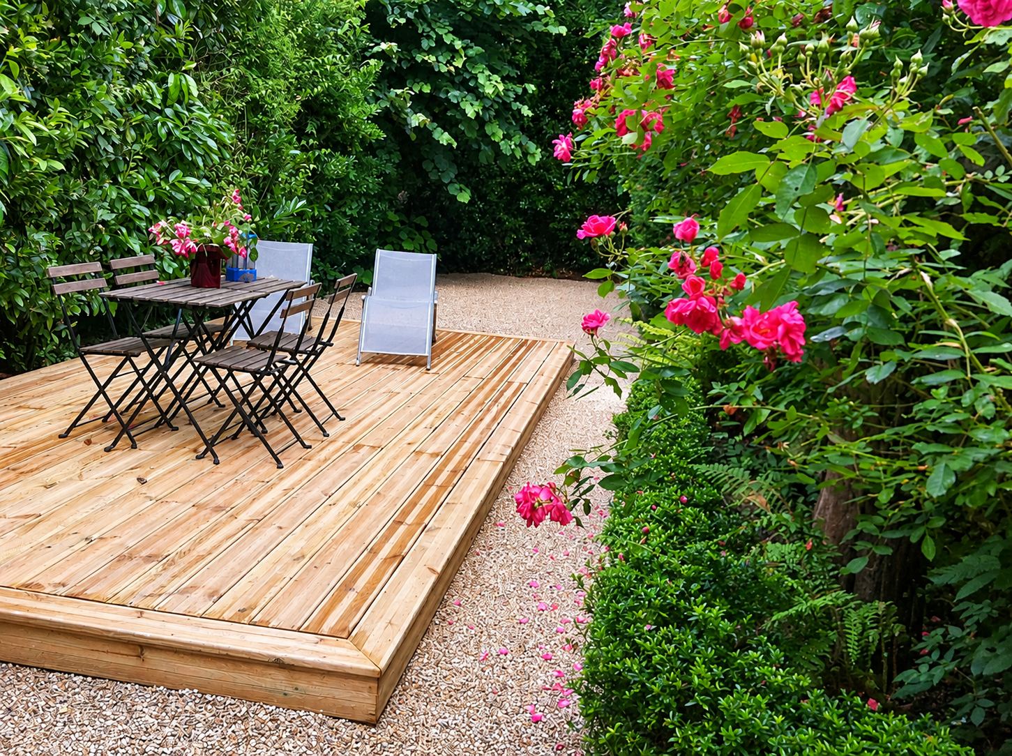 Une terrasse en bois posée sur du gravier dans un jardin, avec une table, des chaises pliantes, des transats et des rosiers en fleurs.